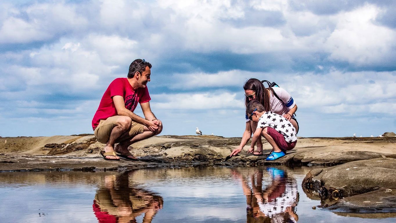 Rob crouching at a tidal pool with his family, their reflections in the still water.